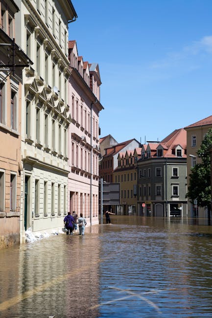 A flooded residential street in Kennington with water covering the road surface and partially submerging parked cars. The scene features trees with green and orange leaves leaning over the flooded are