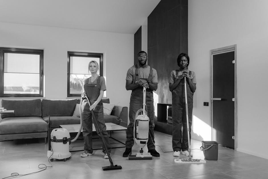 A team of three professional cleaners from cleanersse11.co.uk is conducting surface cleaning and deep cleaning in a modern residential living room. The cleaners are dressed in uniform and equipped wit